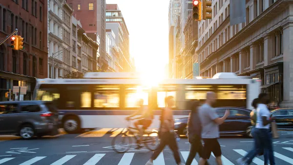 People, cars, bikes and buses traveling through a busy intersection on 5th Avenue and 23rd Street in New York City with shining sunlight background.