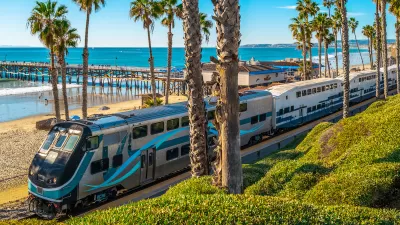 Silver Metrolink commuter train on tracks next to beach in San Clemente, California with palm trees and wooden pier in background.
