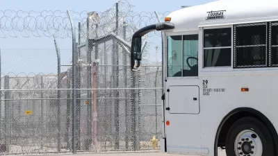 White prison transport bus passing in front of heavily fortified prison fence with barbed wire.