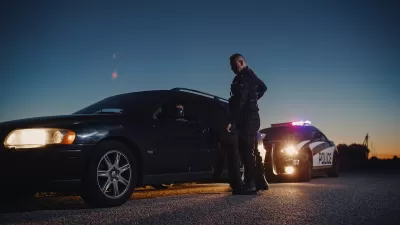 Police office standing next to sedan he just pulled over at night with police car in background.