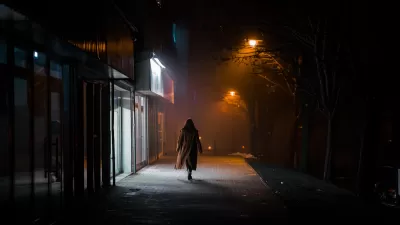 Silhouette of woman in long coat walking on darkened city street alone at night, passing brightly lit sotrefront.