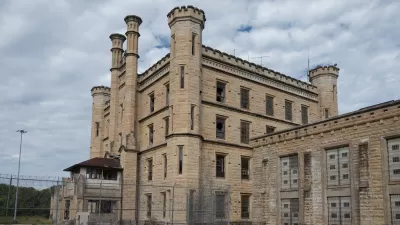 Deralict stone prison building with turrets and a chain link fence in foreground, under a gray, cloudy sky.
