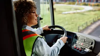 Woman bus driver sitting behind wheel of bus wearing long-sleeved shirt and yellow safety vest.