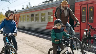 Man and two children on bikes next to red and silver train on sunny day.