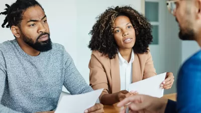 A Black couple holding contracts sits across the desk from a white mortgage lender.
