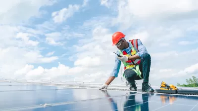Worker wearing orange hard hat and vest over light blue long-sleeved shirt and jeans inspects rooftop solar panel