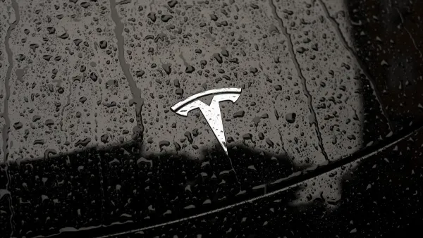 Close-up on silver Tesla logo on black car hood dotted with raindrops.