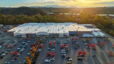 Aerial view of large parking lot in front of large grocery or big-box store with small fall-colored trees among parked cars and sunset sky in background.