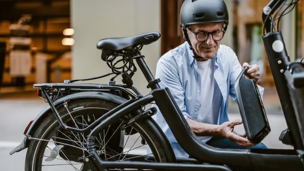 Man wearing black bike helmet changing battery on black e-bike outdoors.