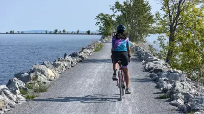 View from behind of woman wearing helmet on bicycle riding on concrete causeway with water on either side.