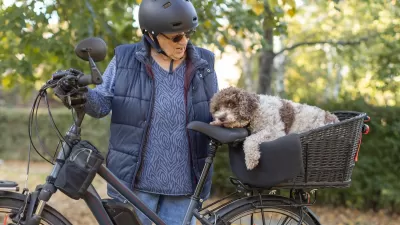 ELderly woman wearing black helmet and blue puffy vest stands next to e-bike with white and brown dog in rear basket.