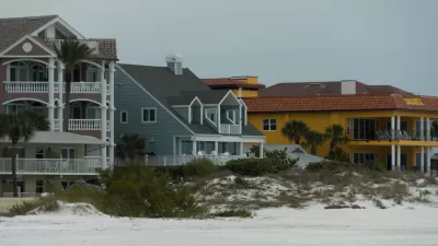 Three colorful, large beachfront homes, one khaki, one blue, and one yellow, with a small dune in front and flat sand in foreground.