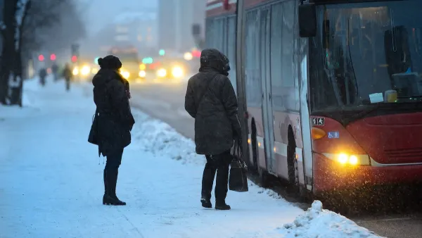 Two people waiting to board arriving bus on a snowy day.