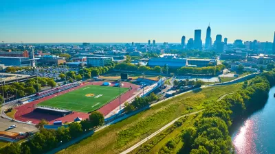 Aerial view of a bright green turf soccer stadium surrounded by red surfaced track with a river to the right and a downown skyline in the background against a bright blue sky.