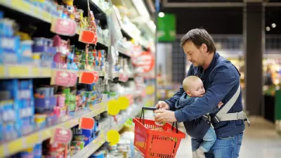 Man with baby in sling holds red grocery basket while looking at refrigerated store section.