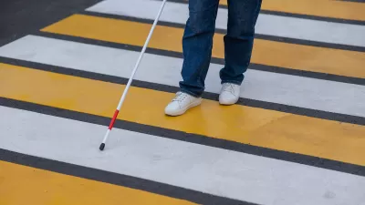 Close-up of person with vision impairment wearing jeans and white sneakers crossing yellow and white striped crosswalk with cane.