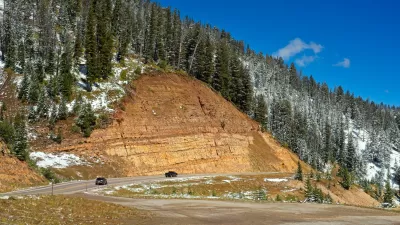 A road winds along the edge of a mountain, with an inclined tan rockface above it and a snowy treeline near the top of a photo.