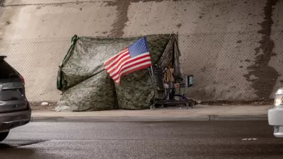 Tent covered with camouflage tarp with American flag on front under freeway overpass in California.