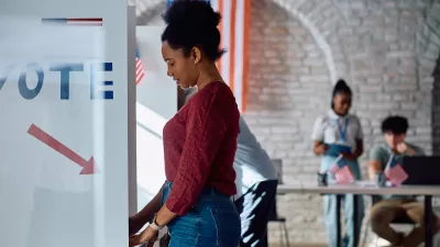 A side view of a young Black woman in a red sweater and jeans standing at a ballot box in a gymnasium with white walls and an American flag in the background.