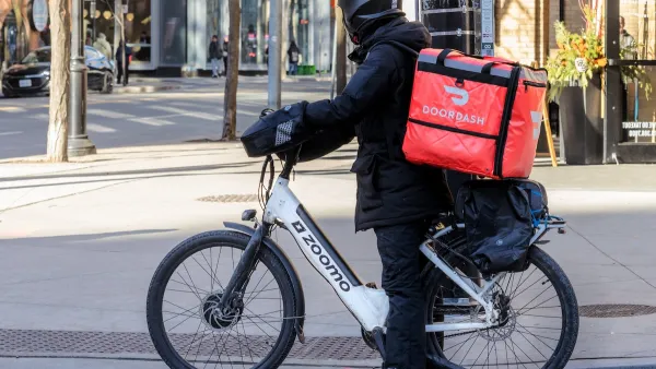 Person in black clothing and helmet with red DoorDash box on back of electric bike on urban street.