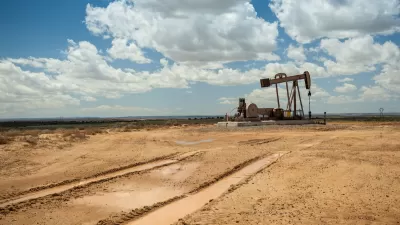 Distant view of brown oil pump jack in a flat desert with cloudy blue sky.