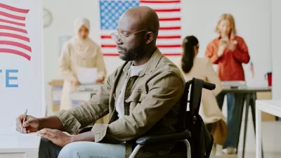 African American man in a wheelchair filling out paper ballot at a polling station with American flags in background.