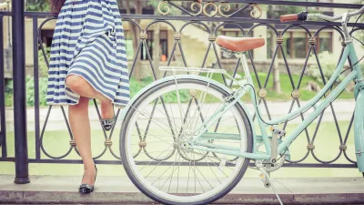 Close-up on woman in white and blue striped knee-length dress standing next to mint green cruiser bike resting against low wrought iron fence in front of green lawn.