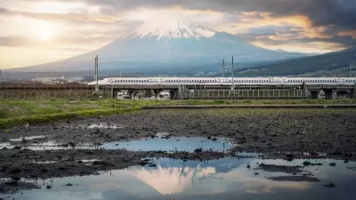 White Shinkansen high-speed rail train passing through green fields with water and Mount Fuji in background in Japan.