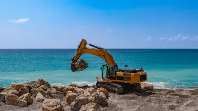 Yellow excavator on rocky beach with bright blue water in background.