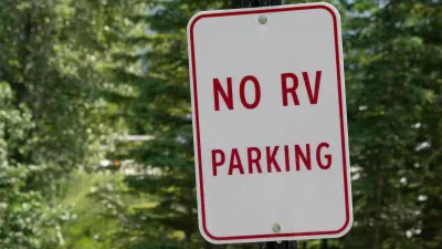 Close-up of white sign with red text 'No RV parking' against green foliage.