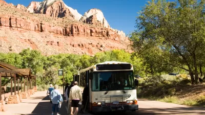 White shuttle bus picks up passengers in Zion National Park, red cliffs and mountains in background.