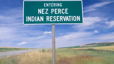 Green roadside sign with white text reading "Entering Nez Perce Indian Reservation" against grassy field and blue sky.