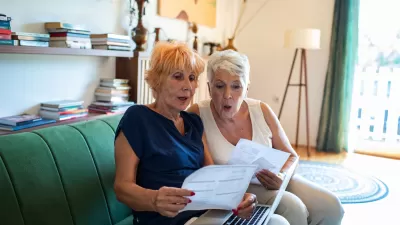 Two older women sitting on green sofa looking at paperwork or bills.