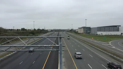 A wide highway with a few cars on a gloomy day. Low-rise commercial buildings and more car infrastructure is visible to the side of the road.