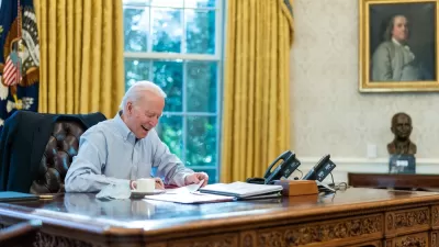 President Jose Biden appears in a collared, button down shirt while signing papers while seated at the desk in the Oval Office of the White House