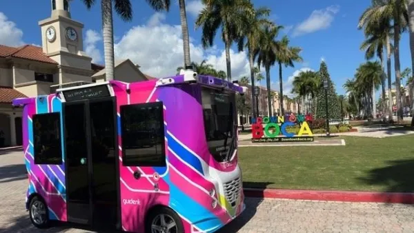 Colorful pink and purple small autonomous transit shuttle in park in Boca Raton, Florida with tall palm trees lining lawn in background.