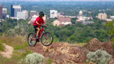 An adolescent boy catches some air on a mountain bike on a hill above the city of Boise. The city’s downtown is clearly visible in the background.