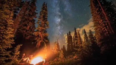 A campfire in the wilderness, surrounded by a group of people, burns with the stars overhead.