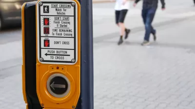 A crosswalk activation button is shown in the foreground with two pedestrians show blurred while crossing the street in the background.
