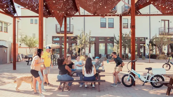 Group of young people sitting at outdoor shaded table laughing and talking with e-bikes and dog next to them.
