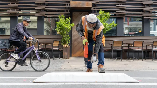 Worker in orange vest installing plastic bike lane separator as man rides past on bicycle in background.