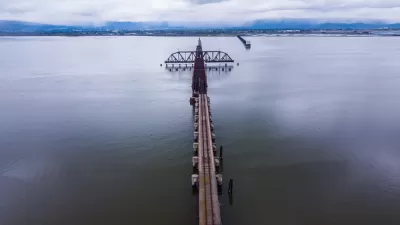 A long rail bridge, rusted and old, crosses a long stretch of water with hills and clouds on the other side.