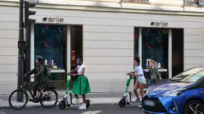A woman on an electric bike and two women on electric scooters wait at an intersection for the light to change.