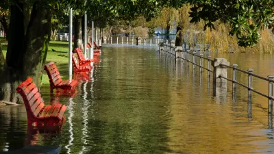 Flood waters overwhelm a park, full of empty benches, on a sunny day.