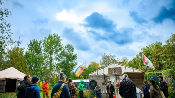 A group of people circle around in front of the Heron’s Nest greenhouse entrance. Two flags ever in the wind as a cloudy sky hovers over the volunteers of the space.