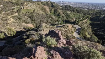 The Griffith Observatory is perched on a hill above Los Angeles, with the city's downtown shown int he distant background. Griffith Park's rolling hills and winding roads are shown in the foreground.