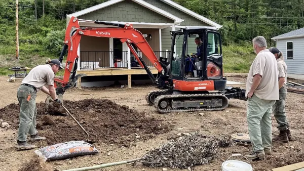 Small green house on unfinished lot with red earthmover and workers in front yard.