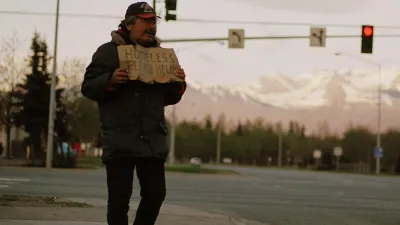 Unhoused man holds up cardboard sign on street corner in Anchorage, Alaska with snowy mountains in background.