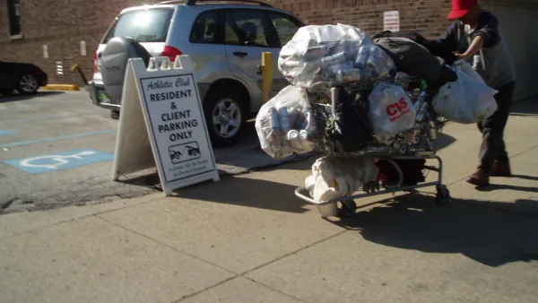 Unhoused man wearing a red bucket hat pushing a shopping cart full of plastic bags full of empty cans.