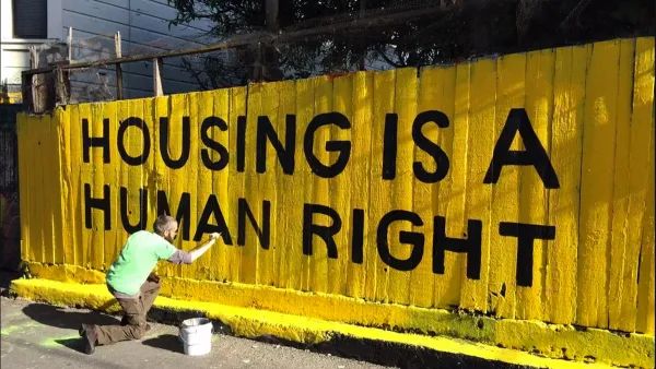 Man in green shirt painting yellow and black "Housing is a human right" mural on fence.